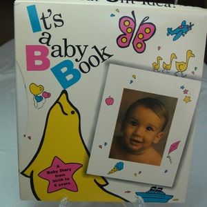 May include: A pink and blue book with the title "It's a Baby Book" and a picture of a baby in a white frame. The book has a pink star with the text "A Baby Diary from birth to 5 years".