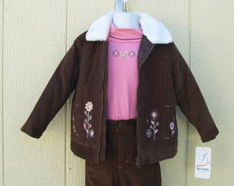 Op de afbeelding: Bruine corduroy jas met een nep bontkraag en bloemenborduursel. De jas wordt gecombineerd met een bruine corduroy broek en een roze turtleneck.