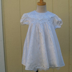 Puede incluir: Vestido de bebé de satén blanco con cuello y ribete de encaje. El vestido tiene un corpiño fruncido y una falda completa.