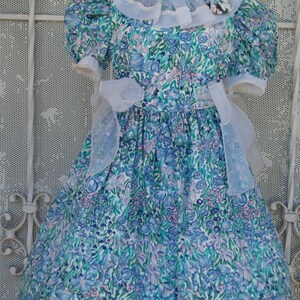 May include: A blue and white floral dress with a white lace collar and a sheer blue bow. The dress has puffed sleeves and a full skirt.