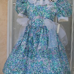 May include: A white dress with a blue and green floral print. The dress has a white lace collar and a white sash. The dress is on a mannequin.