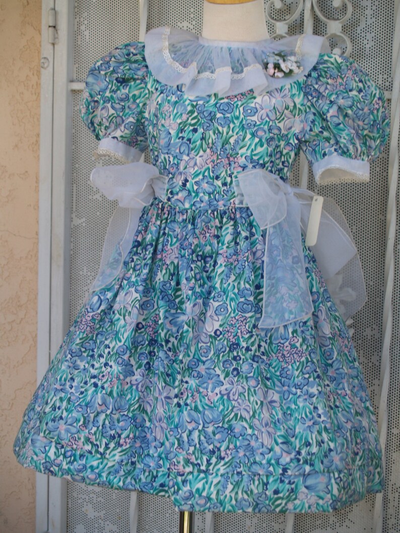 May include: A white dress with a blue and green floral print. The dress has a white lace collar and a white sash. The dress is on a mannequin.