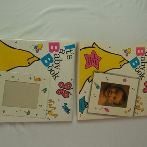 May include: Two yellow and white baby books with a dolphin design. The books have a photo frame on the front cover. The text on the cover reads "It's Baby Book".