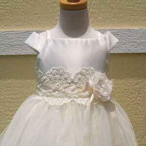 May include: A white satin and tulle dress with a lace bodice and a flower accent. The dress has a scoop neckline and cap sleeves.