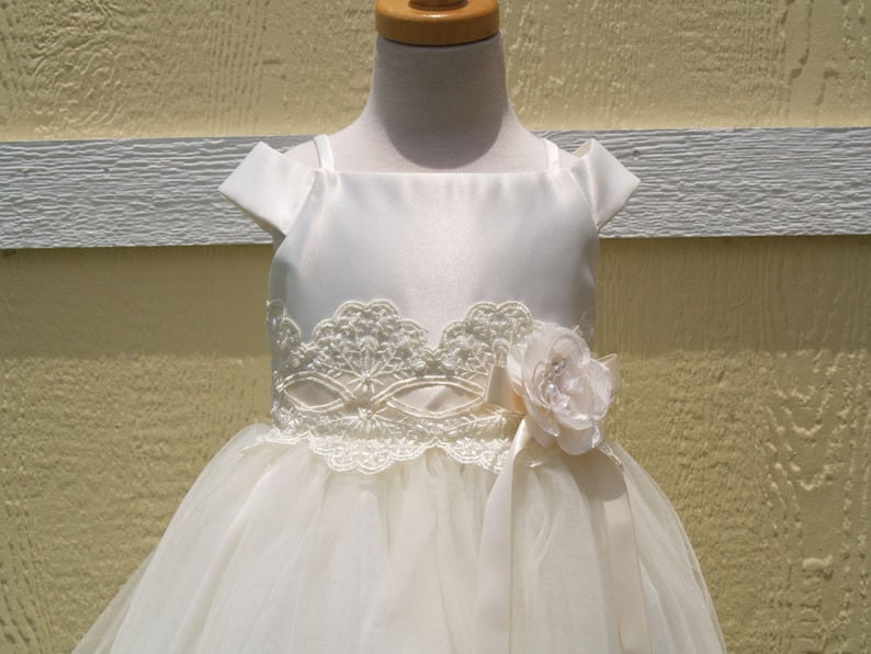 May include: A white satin and tulle dress with a lace bodice and a flower accent. The dress has a scoop neckline and cap sleeves.