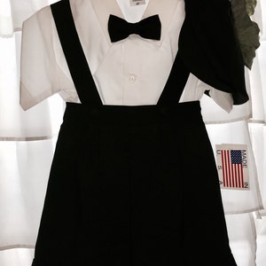 May include: A black and white outfit for a young boy, consisting of a white button-down shirt with a black bow tie, black shorts with suspenders, and a black jacket. The outfit is made in the USA.