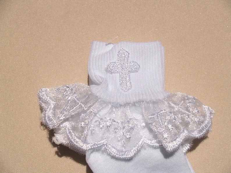 May include: White baby socks with a lace trim and a silver cross embroidered on the top of the sock.