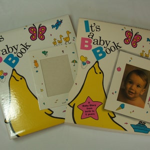 May include: Two baby books featuring a yellow dolphin design and the text "It's a Baby Book". The books include photo frames and are decorated with colourful butterflies, aeroplanes, and ducks.