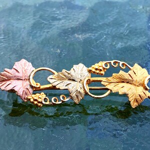 Puede incluir: Un broche de tono dorado con un diseño de vid. Las hojas están hechas de diferentes tonos de oro y las uvas están hechas de pequeñas cuentas doradas.