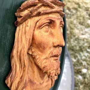 May include: A wooden sculpture of a man's face with a crown of thorns. The sculpture is painted in shades of brown and beige and is mounted on a green background.