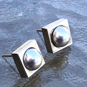 May include: A pair of silver earrings with a round pearl-like center piece set in a square frame.