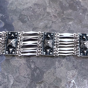 May include: Silver bracelet with a repeating pattern of square black and silver decorative elements. The bracelet has a series of silver bars connecting the decorative squares. The bracelet is designed with a clasp closure.