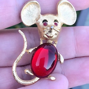 Op de afbeelding: Een gouden muis broche met een grote rode cabochon steen. De muis heeft een glimlachend gezicht met rode ogen en een lange, gekrulde staart.