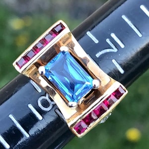 Könnte beinhalten: Ein goldener Ring mit einem blauen Edelstein und roten Rubin-Akzenten. Der Ring ist in einer quadratischen Form mit einem geschwungenen Design gefasst.