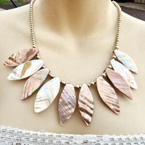 May include: A necklace featuring iridescent, leaf-shaped pendants in shades of pink, white, and brown. The pendants are strung on a beaded gold-colored chain. The necklace is displayed on a mannequin.