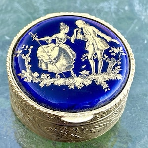 Può includere: Un piccolo portagioie rotondo e antico con esterno in metallo color oro. Il coperchio presenta una superficie in smalto blu scuro con un'illustrazione dorata di due figure in una scena romantica. La scatola ha un design decorativo e strutturato.
