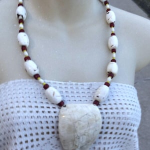 May include: A white shell necklace with red and yellow beads. The necklace has a large, white shell pendant with a carved design.