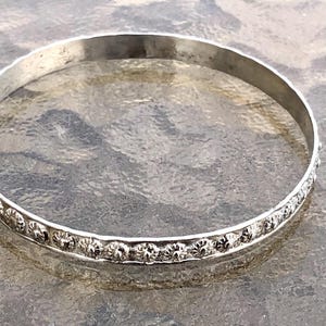 Peut inclure: Un bracelet jonc de couleur argentée. Le bracelet a un design simple et rond avec une série de petits motifs floraux répétitifs sur le bord extérieur. Le bracelet est présenté sur une surface texturée.