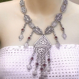May include: A silver necklace with a large, ornate pendant and dangling red stones. The pendant is shaped like a diamond and has intricate filigree designs. The necklace is made of metal and has a delicate chain.