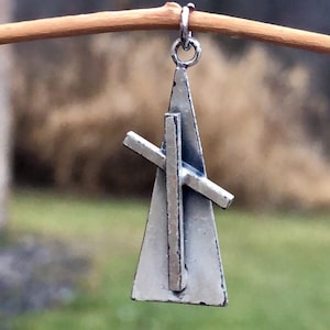 May include: A silver pendant charm in the shape of a triangle with a cross inside. The pendant is hanging from a twig.