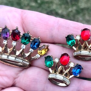 May include: A set of two gold-tone crown-shaped brooches with colorful rhinestones. The larger brooch has seven rhinestones in shades of green, purple, red, and blue. The smaller brooch has three rhinestones in shades of green, red, and blue.