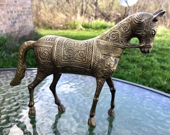 May include: A gold-colored metal horse figurine with intricate floral designs. The horse is standing on a glass table.