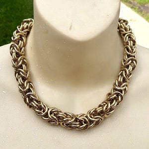 Puede incluir: Un collar de cadena bizantina en tono dorado. El collar se exhibe en un maniquí. La cadena está hecha de bucles entrelazados, creando una apariencia texturizada y ornamentada. El collar es una pieza llamativa, adecuada para diversas ocasiones.
