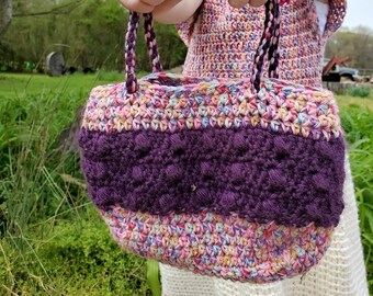 Könnte beinhalten: Ein junges Mädchen hält eine bunte gehäkelte Tragetasche mit einem violetten Streifen. Die Tasche hat einen weißen gehäkelten Riemen.
