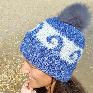 May include: A blue and white knitted beanie with a fluffy grey pom pom on top. The beanie is being worn by a person with long brown hair.