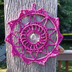 Può includere: Un acchiappasogni all'uncinetto magenta vibrante con una perla di cristallo centrale. Il design circolare presenta intricati motivi traforati e un bordo smerlato. È appeso a una superficie di legno, mostrando uno stile bohémien fatto a mano.