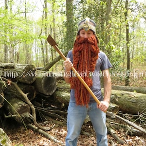 Peut inclure: Une personne portant un chapeau tricoté bleu et marron avec une longue barbe tricotée orange et marron, tenant une hache en bois dans un décor forestier.