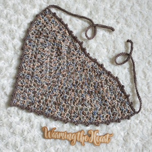 May include: A crocheted triangle top with brown, beige, and blue yarn. The top has two long ties that can be tied around the neck. The top is on a white fluffy surface. The text "Warming the Heart" is written on a wooden sign.