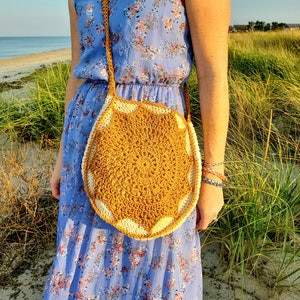 May include: A crocheted mustard yellow and white circular crossbody bag with a long strap. The bag is worn by a person wearing a blue floral dress.