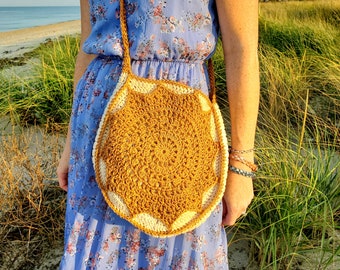 Può includere: Una borsa a tracolla rotonda in crochet giallo senape e bianco con un cinturino lungo. La borsa è indossata da una persona che indossa un abito floreale blu.