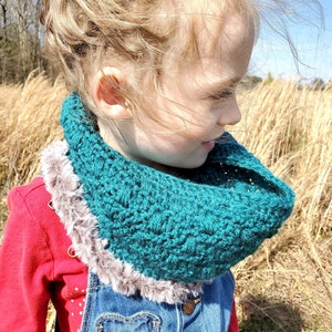 Può includere: Una bambina indossa una sciarpa infinita all'uncinetto color turchese con un bordo in pelliccia sintetica marrone. Indossa una camicia rossa e una salopette blu con una tasca a forma di cuore.