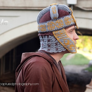Puede incluir: Una persona lleva un gorro de punto gris y marrón con ribete amarillo y marrón. El gorro tiene un diseño único que se asemeja a un casco. La persona lleva una chaqueta marrón con capucha.
