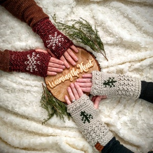 May include: A cozy image featuring four pairs of knitted fingerless gloves in various colors. Two pairs are burgundy with white snowflake patterns, and two are beige with green tree designs. The gloves are arranged around a wooden disc with the text "Warming the Heart".