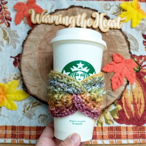 May include: A white coffee cup with a green Starbucks logo is wrapped in a colourful crocheted cosy. The cosy is made with a variety of colours, including red, orange, yellow, and green. The cup is sitting on a wooden surface with a sign that says "Warming the Heart".