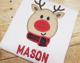 May include: White t-shirt with a red and brown embroidered reindeer with a red nose and a red and black plaid scarf. The name "MASON" is embroidered in red below the reindeer.