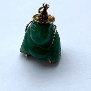 Peut inclure: Un pendentif en jade vert représentant une figure assise portant un chapeau en or avec une pierre précieuse rouge. Le pendentif est sur un fond blanc.