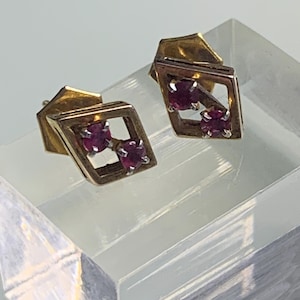 May include: A pair of gold stud earrings, each featuring a geometric diamond-shaped frame. Within each frame, two round, deep pinkish-red gemstones are set in silver-toned prongs, creating a balanced and structured appearance for these jewelry pieces.