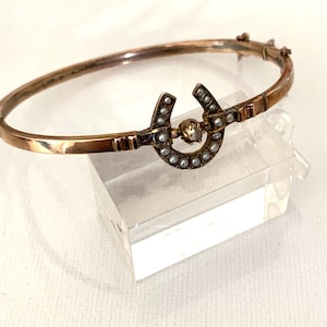 Könnte beinhalten: Ein goldenes Armreif-Armband mit einem hufeisenförmigen Anhänger, der mit Perlen besetzt ist. Der Anhänger hat eine einzelne, größere Perle in der Mitte.
