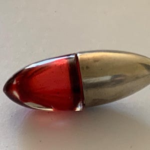 Peut inclure: Un petit flacon de parfum ovale, composé d'une section en verre transparent remplie de liquide rouge et d'un capuchon en métal argenté. Le flacon est divisé en deux parties distinctes.