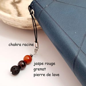Peut inclure: Un cordon noir avec un charme argenté attaché. Le charme a trois perles : une perle de lave brun foncé, une perle de grenat rouge foncé et une perle de jaspe rouge. Le texte "chakra racine", "jaspe rouge", "grenat" et "pierre de lave" est écrit sous le charme.