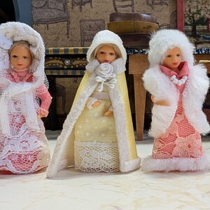 May include: Three miniature dolls standing side by side. The left doll wears a pink dress with a white lace overlay and bonnet. The center doll wears a yellow dress with a white hooded cape. The right doll wears a pink lace dress with a white faux fur vest.