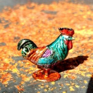May include: A small, decorative rooster figurine featuring a vibrant, multi-colored enamel finish. The bird displays a bright red comb and wattle, with a body patterned in shades of teal, green, purple, and metallic copper, standing on a circular base.