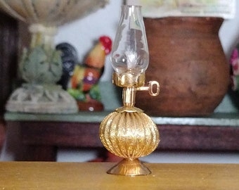 May include: A gold-colored oil lamp with a clear glass chimney and a decorative, ribbed base. The lamp is sitting on a wooden surface.
