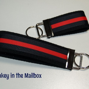 May include: Two black keychains with a red stripe and silver key rings. The keychains are made of woven fabric.