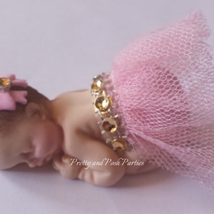 May include: A small, sleeping baby figurine made of fondant, wearing a pink tulle tutu and a pink bow with a gold rhinestone. The figurine is decorated with a gold rhinestone trim.