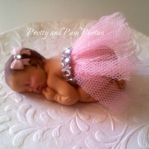 May include: A sleeping baby doll cake topper with a pink tulle skirt and a pink bow with a rhinestone in the center. The doll is lying on a white cake plate with a floral design.